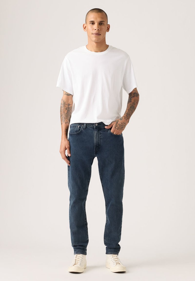 Balti trumpomis rankovėmis marškinėliai, mėlynos spalvos džinsai iš džinso audinio ir smėlio spalvos sportbačiai. Džinsai turi klasikinį kirpimą, džinso tekstūrą ir subtilų siuvimą.