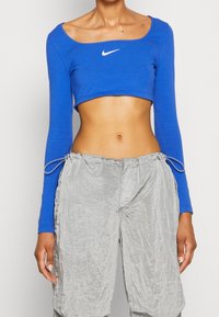 Blå croppad långärmad topp med rund halsringning och vit Nike-logotyp; ihop med ljusgrå, texturerade, löst sittande byxor med snören.