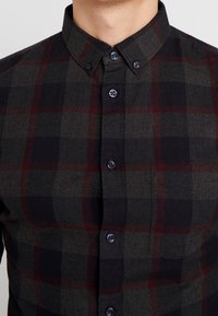 Camicia a quadri scura nelle tonalità di grigio, nero e bordeaux. Presenta un colletto a vento, una piccola tasca sul petto e bottoni scuri. Tessuto morbido.