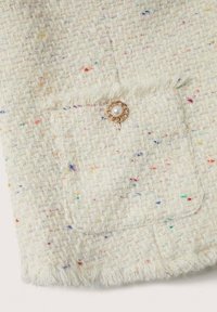 Petite poche en tweed avec bords à franges sur un tissu clair avec des taches multicolores et un bouton rond doré décoratif avec un centre en perle.