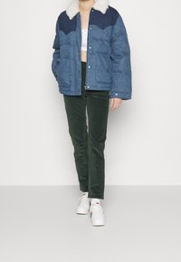 Personne portant une veste en duvet bleue avec un col en polaire blanc, un crop top blanc, un pantalon vert foncé et des baskets blanches se tenant contre un fond uni.