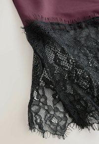 Melna ziedu mežģīņu audums pārklāts pār tumši mauvas krāsas drānu uz gaiša fona, rādot izsmalcinātas rakstu detaļas un smalkas bārkstis.