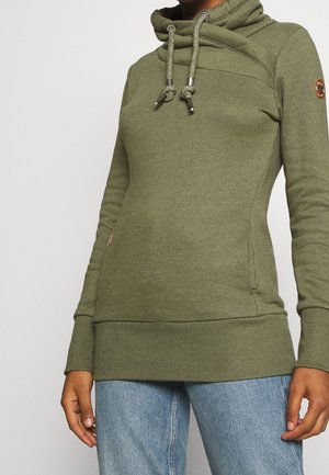 Zbliżenie osoby noszącej oliwkowozieloną bluzę z kapturem i sznureczkami oraz jasnoniebieskie dżinsy, stojącej na jednolitym tle.