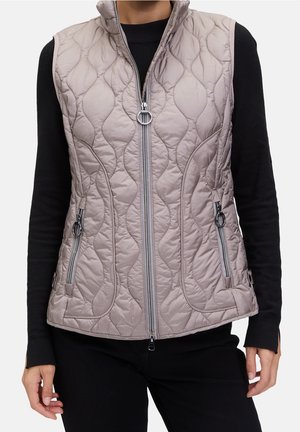 Femme portant une veste matelassée taupe avec fermeture éclair à l'avant et poches zippées par-dessus une chemise noire à manches longues et un pantalon noir.