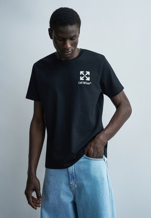 Άνδρας που φοράει μαύρο t-shirt Off-White με λευκό λογότυπο στο στήθος, ανοιχτό μπλε τζην, κοιτάζει προς τα κάτω με το ένα χέρι στην τσέπη.