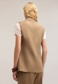 Blazer tanpa lengan berwarna coklat muda dengan potongan yang disesuaikan, menampilkan kerah notch dan bagian belakang yang terstruktur. Dipakai di atas atasan lengan pendek.