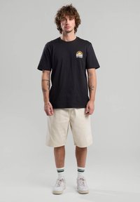 T-shirt nera in cotone con maniche corte, caratterizzata da una piccola grafica gialla sul petto sinistro; abbinata a pantaloni corti crema e sneakers bianche.