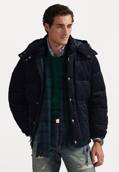 Navy puffer jacket with a corduroy texture, snap closures, and adjustable hood. Worn over a green knit sweater and plaid scarf.