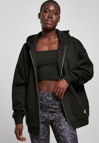 Schwarze Kapuzenjacke mit vollem Reißverschluss, übergroßer Schnitt, gerippte Bündchen und Vordertaschen, kombiniert mit gemusterten Leggings, die florale Motive zeigen.