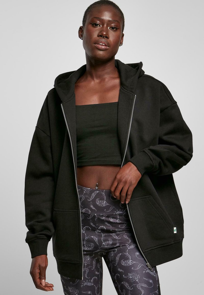 Schwarze Kapuzenjacke mit vollem Reißverschluss, übergroßer Schnitt, gerippte Bündchen und Vordertaschen, kombiniert mit gemusterten Leggings, die florale Motive zeigen.