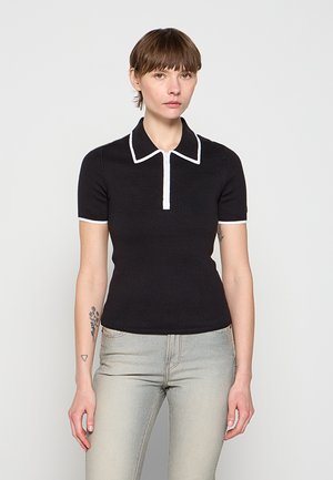 Kobieta ubrana w dopasowany czarny krótki top z białą lamówką na kołnierzyku i rękawach, w połączeniu z jasnoszarymi dżinsami, stojąca na jednolitym tle.