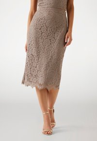 Falda lápiz de encaje en taupe con patrón floral, dobladillo ondulado y un sutil brillo. Combinada con sandalias metálicas de tiras finas.
