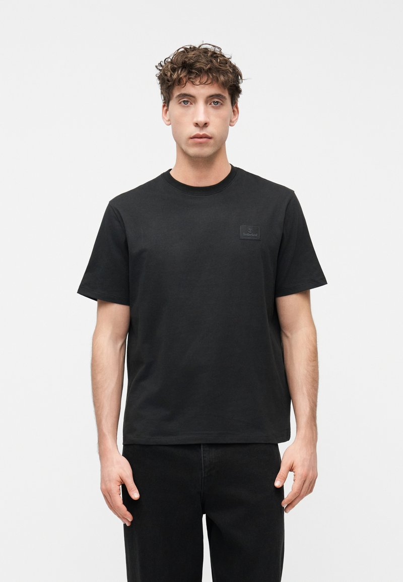 Νεαρός με σγουρά μαλλιά φοράει απλό μαύρο t-shirt Timberland και μαύρο παντελόνι, στέκεται μπροστά σε λευκό φόντο, κοιτάζοντας προς τα εμπρός.