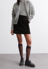 Cardigan grigio a costine, mini gonna in velluto a coste nera, scarpe nere e calze a rete nere fino al ginocchio. Maglietta a maniche lunghe a righe sotto il cardigan.
