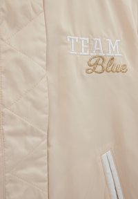 Smėlio spalvos bombonešio striukė su siuvinėta tekstūra, kurioje yra baltos spalvos siuvinėtas žodis „TEAM“ ir auksinės spalvos „Blue“ logotipai, bei balta juostele dekoruota kišenė.