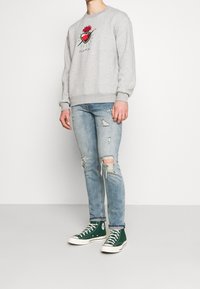 Grå sweatshirt med en röd hjärt- och ros-grafik, ihop med slitna ljusblå jeans och gröna högklackade sneakers.