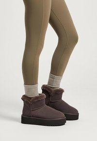 Graue Knöchelstiefel mit einem plüschigen Fellkragen, strukturiertem Obermaterial und einer dickeren schwarzen Plateausohle, getragen mit olivgrünen Leggings und grauen Stricksocken.