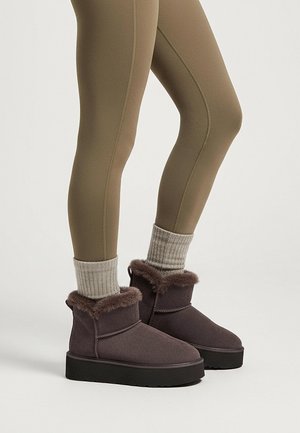 Bottes grises à la cheville avec un col en fausse fourrure, une tige texturée et une épaisse semelle noire, portées avec des leggings vert olive et des chaussettes grises en tricot.