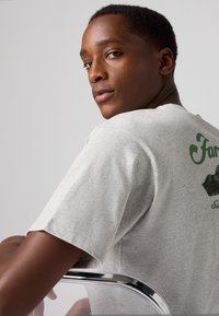 T-shirt en coton gris avec un design graphique vert au dos. Le mannequin a les cheveux courts et est appuyé sur une chaise en chrome.
