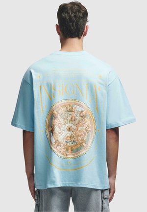 Lyseblå bomulds t-shirt med et stort flerfarvet trykt design på ryggen, med guld accenter og et rundt mønster.
