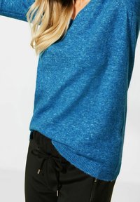 Kvinna i en blå stickad tröja och svarta byxor med dragsnodd, som visar midjan och en del av sitt långa blonda hår.