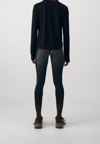 Černý top s dlouhým rukávem v kombinaci se šedými legínami. Legíny mají kontrastní švy a texturované detaily; modelka má na sobě outdoorovou obuv.