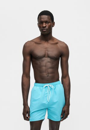 Homme musclé à la peau foncée portant un short de bain bleu vif avec un cordon blanc, debout devant un fond clair uni.