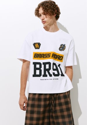 Młody mężczyzna z kręconymi włosami nosi białą koszulkę z grafiką z żółtym i czarnym napisem "BRASS ROAD BR91" oraz brązowe kraciaste spodenki, stoi przy białej ścianie.