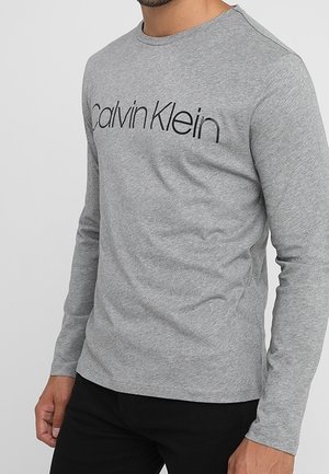 Mężczyzna noszący jasnoszarą koszulę z długim rękawem z logo „Calvin Klein” w czarnym napisie na piersi, w połączeniu z czarnymi spodniami.