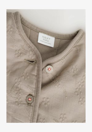 Bež bombažna otroška majica s cvetličnim vezenjem, okroglim izrezom in detajlom s pritisnim gumbom. Oznaka pravi "NEXT baby, izdelano na Šrilanki."