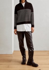 Maglione a righe bianche e nere in maglia con colletto alto, indossato sopra una camicia bianca; abbinato a pantaloni di pelle marrone e stivali neri robusti.