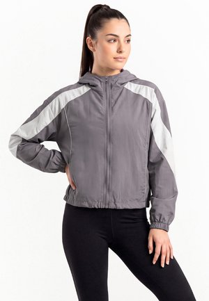 Vrouw met donker haar in een paardenstaart, draagt een grijze capuchontrui met witte strepen op de mouwen en zwarte leggings, staat met één hand op de heup.