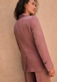 Dopasowana marynarka w jasnym mauve, z wykładanym kołnierzem, jednolitym zapięciem na guzik i subtelnym połyskiem. Tkanina ma gładką teksturę i boczne rozcięcia.