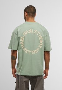 Lysegrønn bomull T-skjorte med korte ermer og løs passform. Baksiden har stor beige sirkeltekst som sier "KARL KANI STUDIOS EST. 1989."