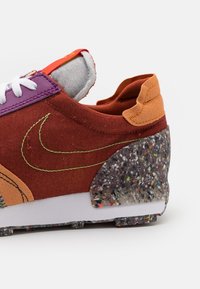 Gros plan d'une basket multicolore montrant un tissu marron, une zone d'œillets violette, une languette de talon orange, des lacets blancs et un support de talon texturé tacheté.