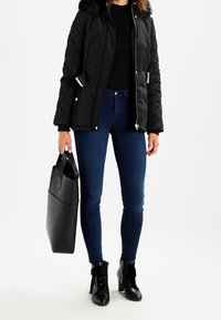 Mustat tikatut takki, jossa on tekoturkiskaulus, yhdistettynä tummaan kapeaan housuun ja nilkkureihin. Toisessa kädessä pidetään teksturoitua mustaa käsilaukkua.