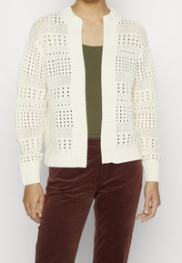 Žena v bílé kardiganu s otevřeným pletením přes olivově zelený top a hnědé kordové kalhoty, stojící na neutrálním pozadí.