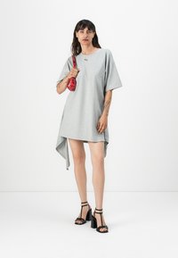Gray, oversized t-shirt dress with an asymmetrical hem. Model holds a small red handbag, wearing black high-heeled sandals.