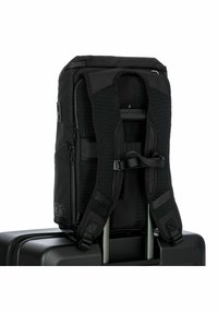 Mochila negra con correas de malla acolchadas, cremalleras y detalles de hardware. Diseñada para ajustarse de forma segura a una maleta con ruedas.