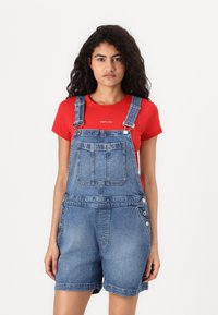 Salopete de ganga com alças ajustáveis, bolso frontal e detalhe de botão na lateral, combinada com uma camisola vermelha.