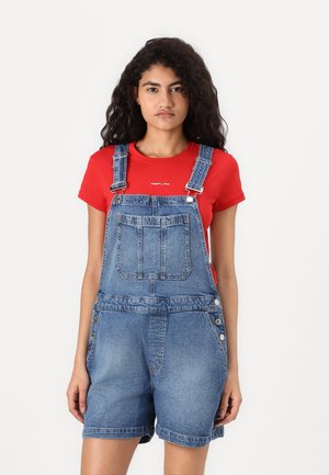 Salopette in denim con spalline regolabili, tasca frontale e dettaglio con bottoni laterali, abbinata a una maglietta rossa.
