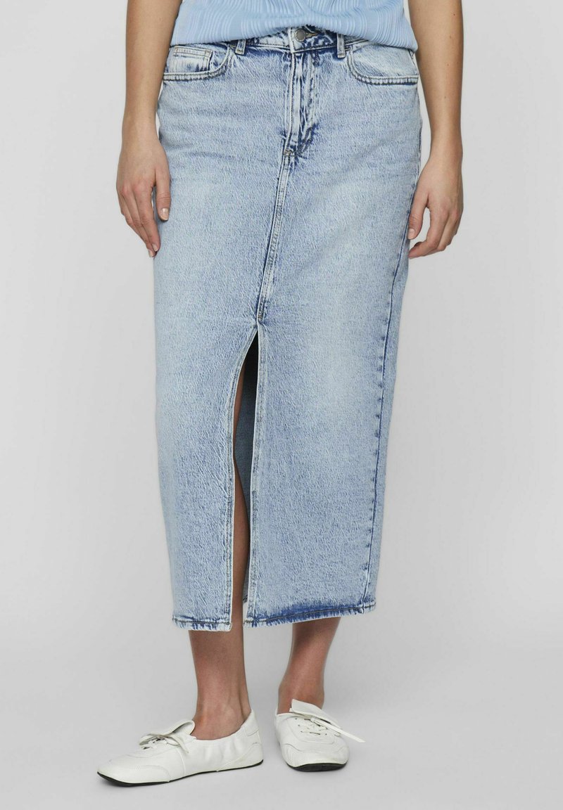 Jupe en denim bleu clair avec une fente à l'avant, longueur cheville et ourlet brut. Portée avec des baskets blanches, mettant en valeur un design décontracté.