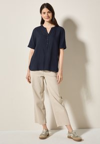 Camicetta blu navy a maniche corte con scollatura a V e bottoni, abbinata a pantaloni larghi beige chiaro e sneaker grigie.