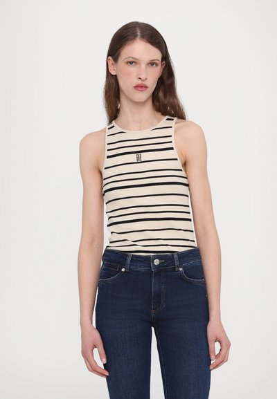 Beige mouwloos topje met zwarte horizontale strepen en een klein logo in het midden. Gecombineerd met donkerblauwe hoge taille jeans.