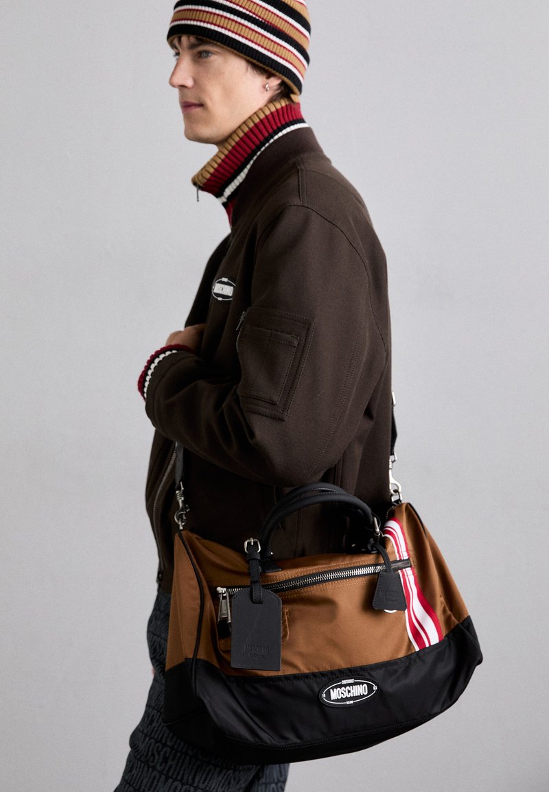 Barna és fekete Moschino táska cipzáros záródással, dupla hordozófüllel és mintás kiegészítővel, amely piros és fehér csíkokat tartalmaz.