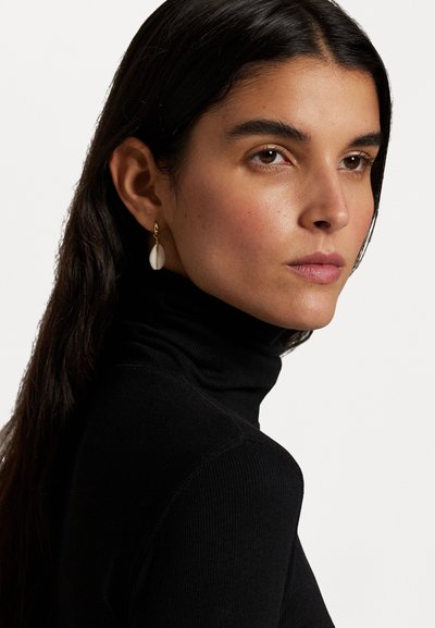 Orecchini a cerchio in oro con un pendente in conchiglia bianca texturizzata, indossati con un top a collo alto nero. Il soggetto ha i capelli lunghi e scuri.