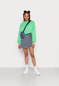 Sweatshirt verde oversize, saia mini padrão azul-marinho, ténis brancos chunky e uma pequena mala de ombro azul-marinho. Cores vivas e texturas em camadas.