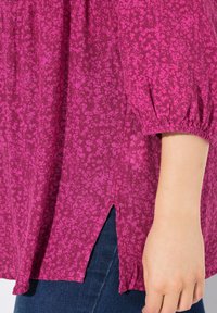 Blusa viola a fantasia con maniche a sbuffo corte, caratterizzata da uno spacco laterale e tessuto testurizzato. Abbinata a jeans blu scuro.