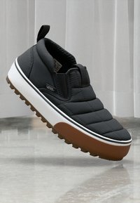 Svarte slip-on sko med en kviltet overdel, hvit mellomsåle og brun gummisåle med teksturert grep. Merkevarelogo synlig på siden.