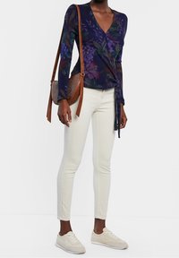 Mujer vistiendo pantalones blancos ajustados, zapatillas crema, blusa de manga larga con estampado floral morado y bolso de hombro de cuero marrón, contra un fondo blanco.